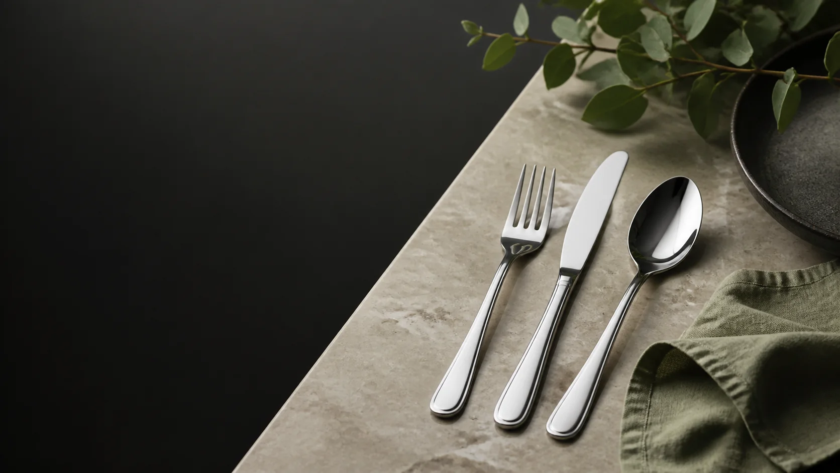 Polished stainless steel cutlery arranged on pale stone with sage linen